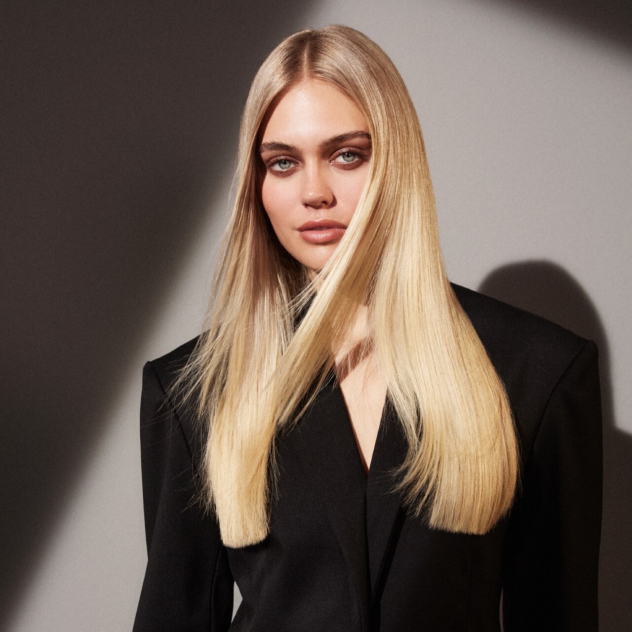 Eine Frau mit langen, glatten blonden Haaren, die einen schwarzen Blazer trägt, steht vor einem grauen Hintergrund mit Farbverlauf und erzeugt einen dramatischen Schatteneffekt.