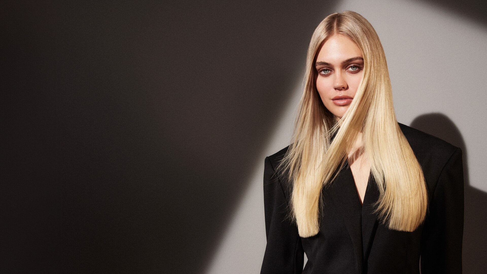 Eine Frau mit langen, glatten blonden Haaren, die einen schwarzen Blazer trägt, steht vor einem grauen Hintergrund mit Farbverlauf und erzeugt einen dramatischen Schatteneffekt.