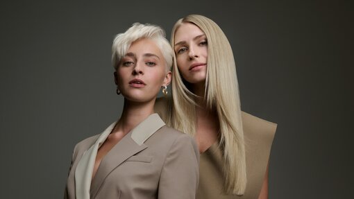 Zwei Models mit blonden Haarvarianten – ein kurzer platinblonder Pixie-Cut und glattes, langes Haar in warmem Blond – präsentieren vielfältige Blondtöne vor einem dunkelgrauen Hintergrund.