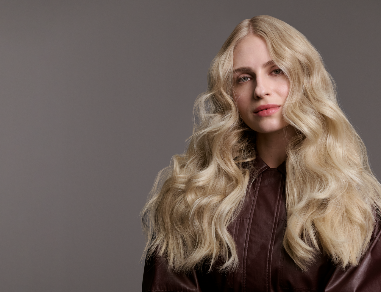 Person mit sehr langem, voluminös welligem, hellblondem Haar trägt eine dunkelbraune Jacke und sitzt vor einfarbig grauem Hintergrund.