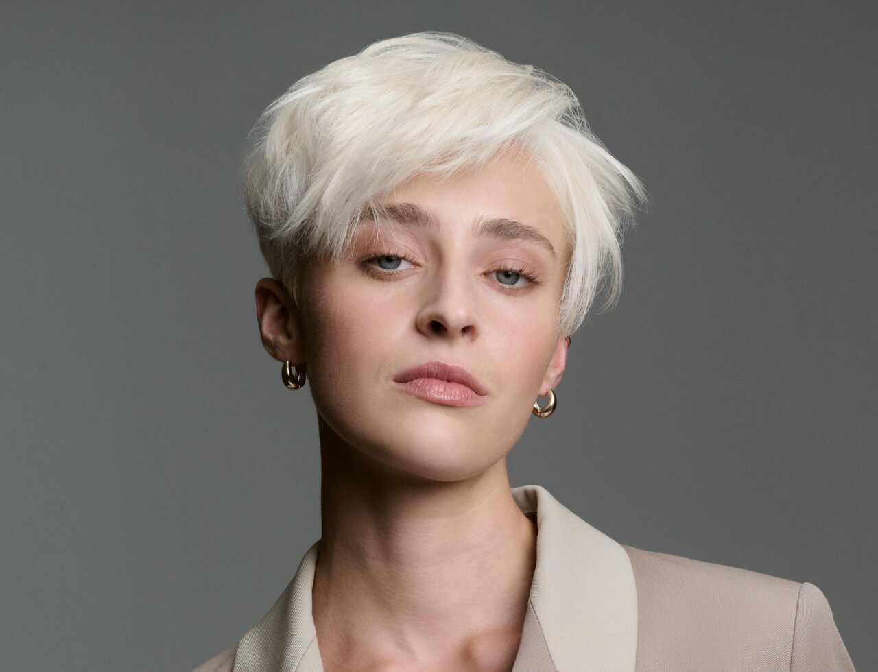 Junge Frau mit eisblonden Haaren, zum kuzren Pixie geschnitten vor grauem Hintergrund.