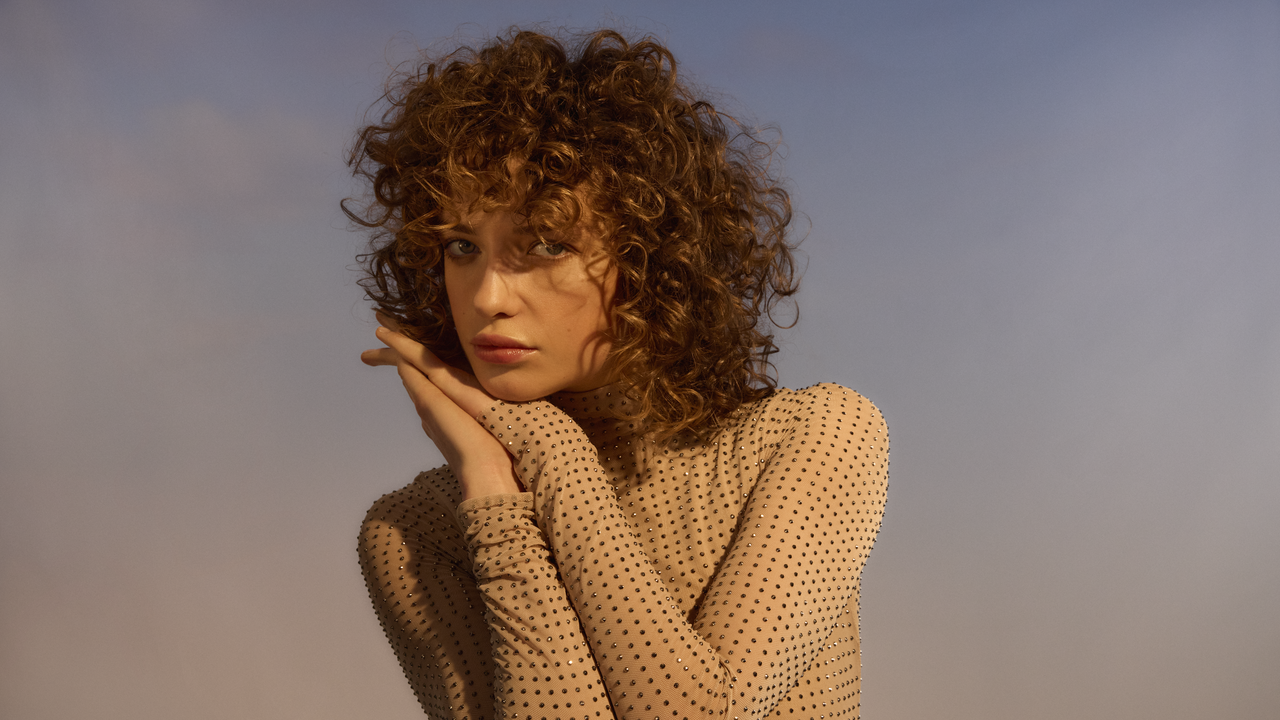 Porträt einer jungen Frau mit schön definierten braunen Locken vor blauem Hintergrund,  sie wird von warmen Licht angestrahlt. 