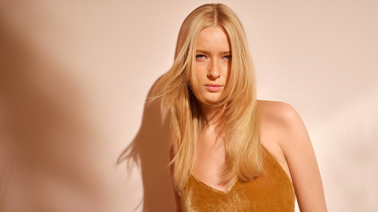 Frau mit mittellangem, leicht gewelltem, hellblondem Haar steht vor warm beige beleuchteter Wand und trägt ein ärmelloses Oberteil in Gold- oder Senfton.