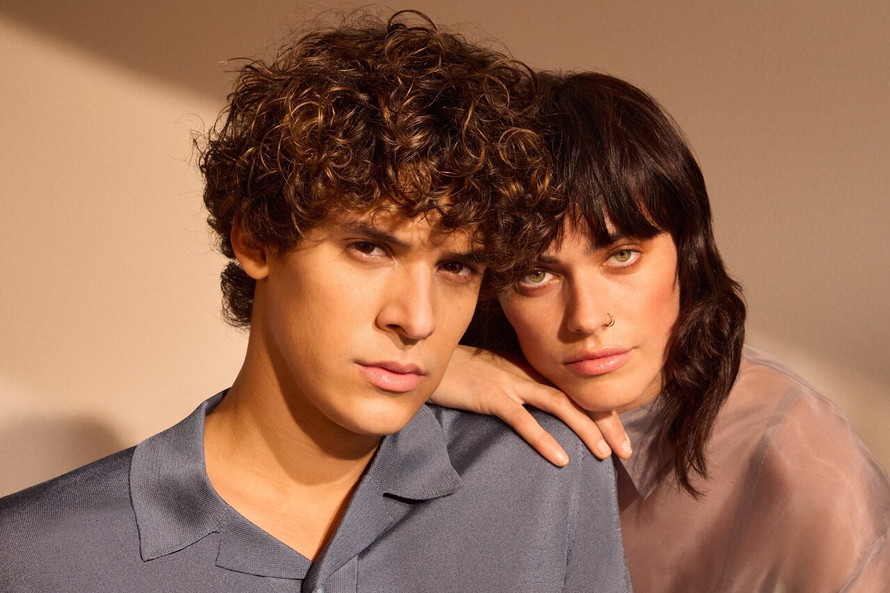 Zwei Personen sitzen eng beieinander. Der junge Mann links hat lockiges kurzes Haar, eine junge Frau rechts mit mittellangem dunklen Haaren; ihre rechte Hand liegt locker auf seiner Schulter.