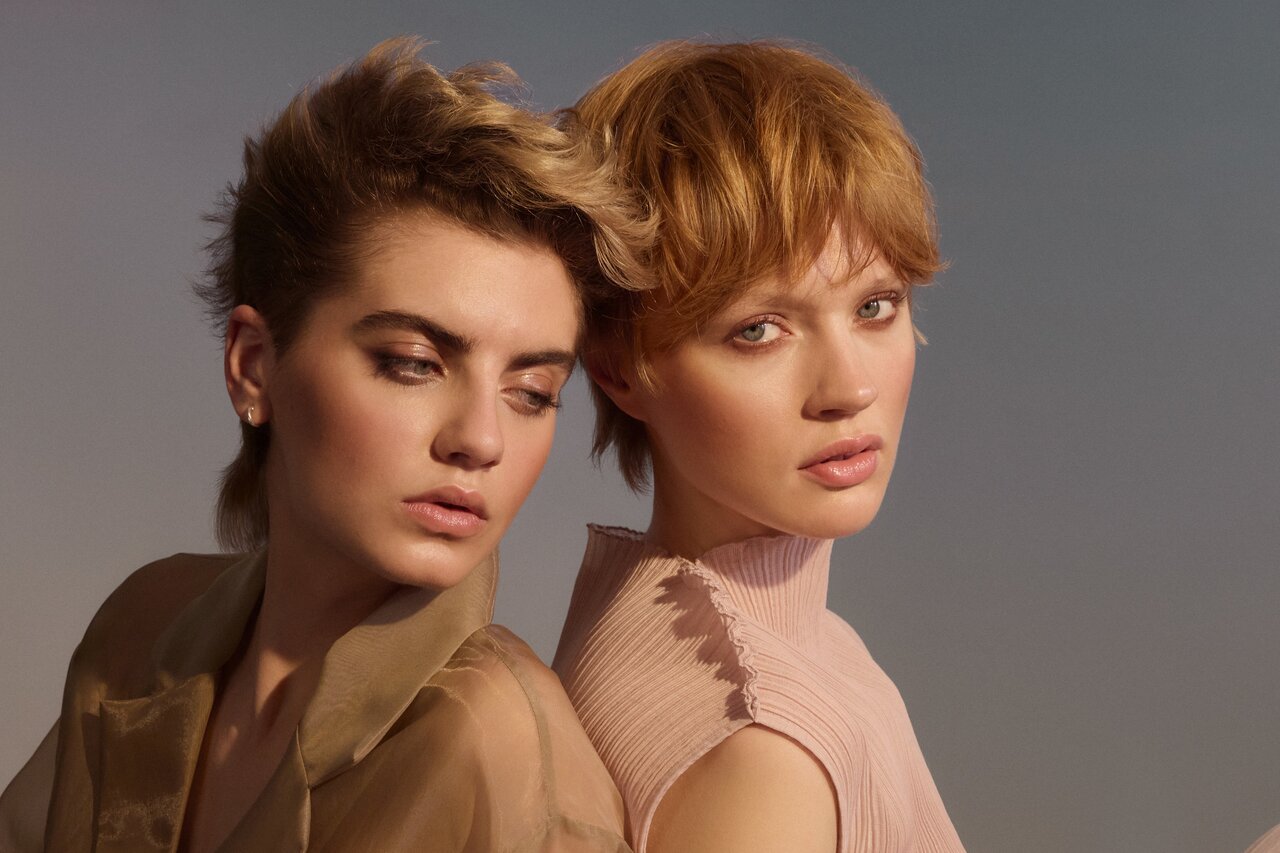 Zwei Personen sitzen Rücken an Rücken vor blauem Himmel; links mit kurz gestyltem, aschblonden Haaren und transparentem Oberteil schaut verträumt aus dem Bild, rechts mit rotblondem Pixie-Cut schaut direkt zum Betrachter.