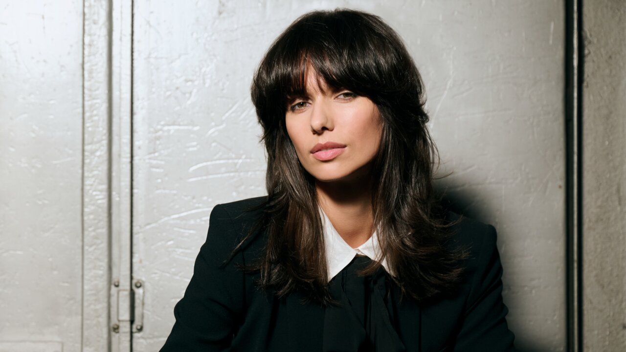 Person mit schulterlangem, voluminösem Haar im dunklen Hosenanzug und weißer Bluse mit Schleife, lässig vor einer metallischen Industrietür sitzend und einen eleganten Business-Look ausstrahlend.