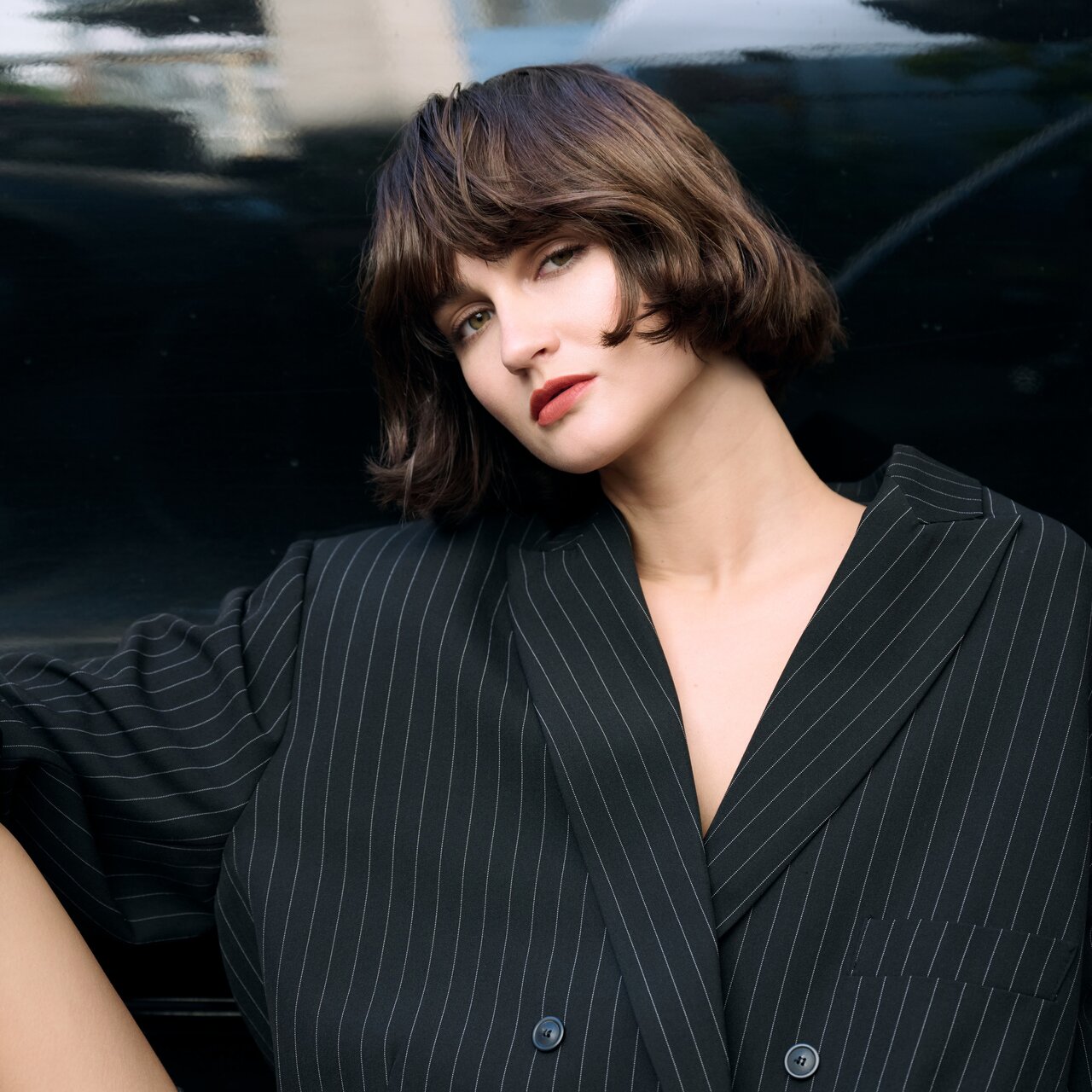 Person mit kinnlangem, glattem Bob in einem oversized geschnittenen, schwarz gestreiften Blazer, lässig vor einer dunklen, spiegelnden Oberfläche posierend und einen modernen, androgynen Streetstyle-Look verkörpernd.