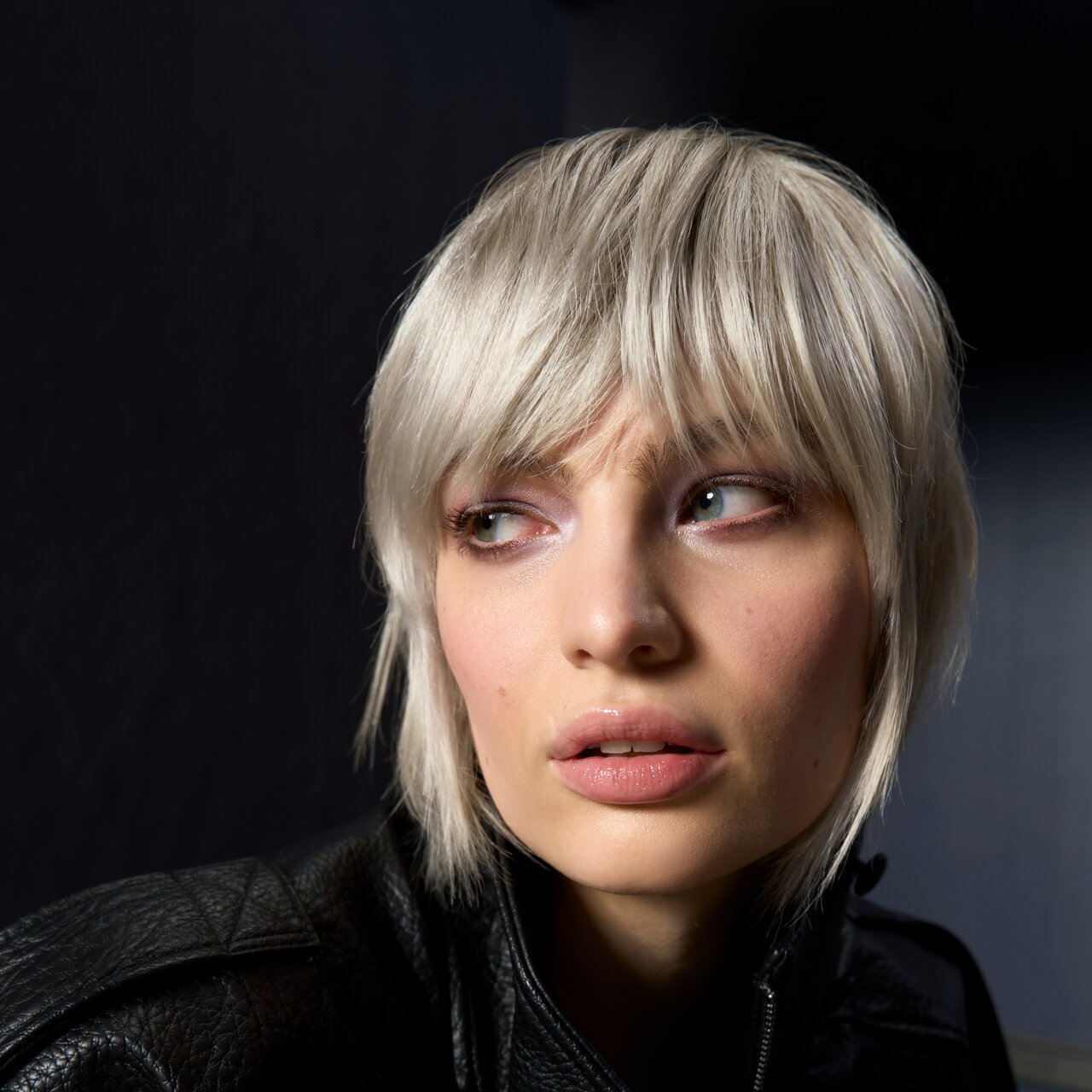 Frau mit platinblondem Bob und Pony in schwarzer Lederjacke vor dunklem Hintergrund