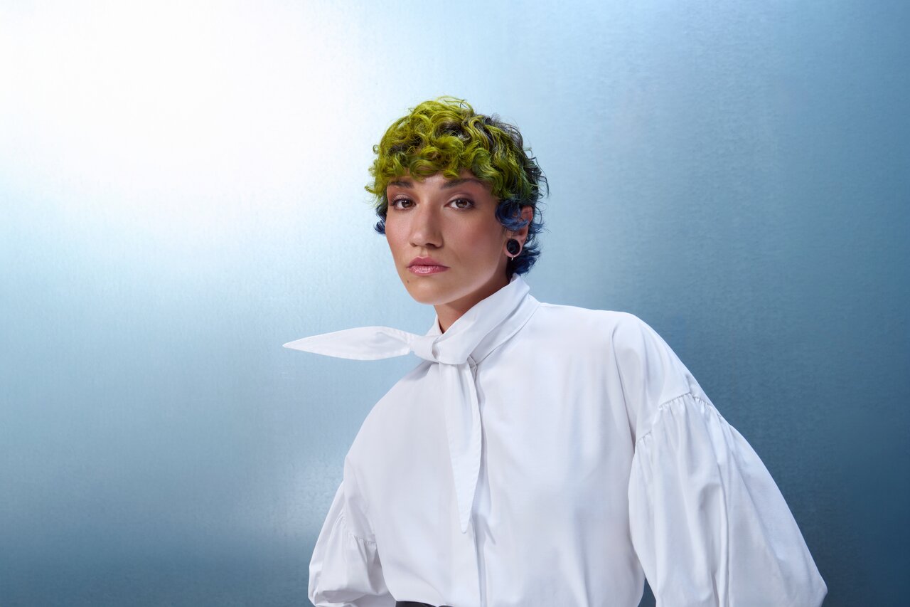 Porträt einer Person mit kurzem, grün gelocktem Haar und weißer Bluse mit Schleifenkragen vor blauem Hintergrund.