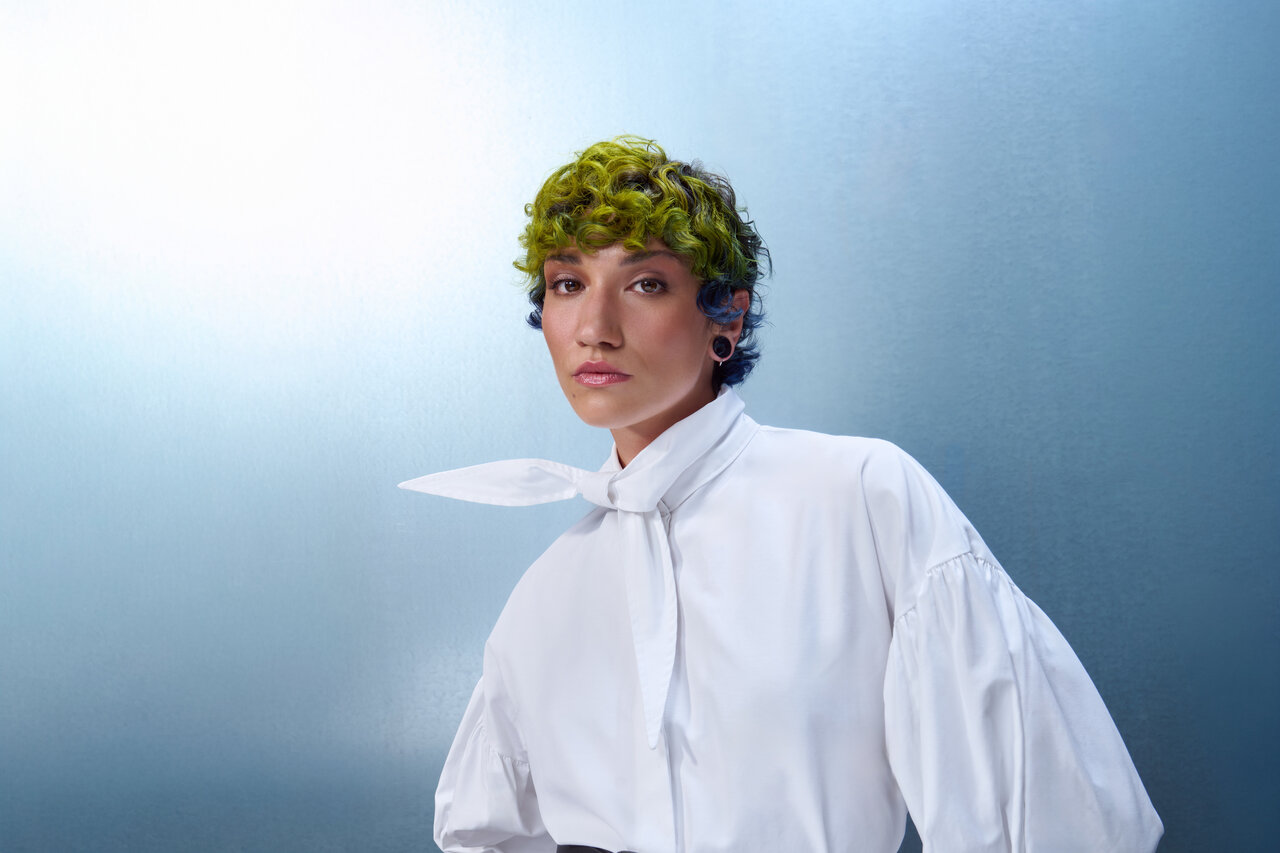 Porträt einer Person mit kurzem, grün gelocktem Haar und weißer Bluse mit Schleifenkragen vor blauem Hintergrund.