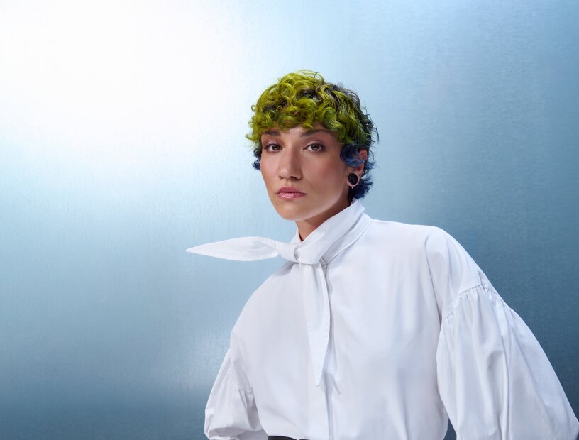 Porträt einer Person mit kurzem, grün gelocktem Haar und weißer Bluse mit Schleifenkragen vor blauem Hintergrund.