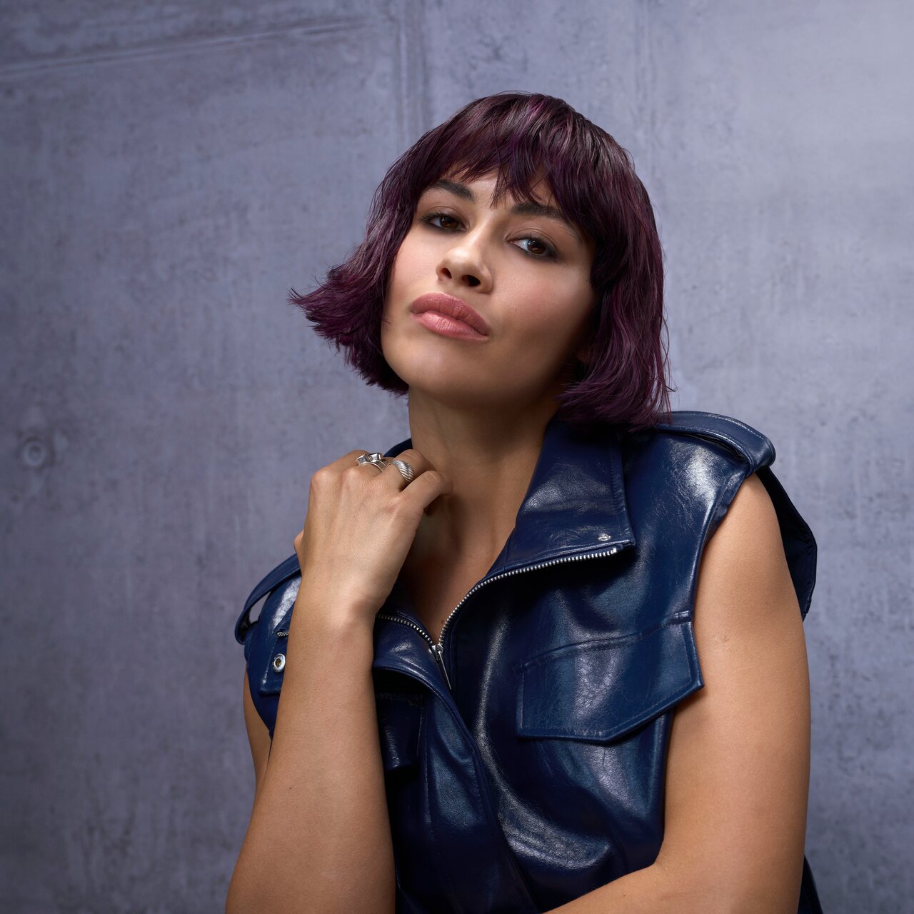 Porträt einer Frau mit kurzem violettem Bob und blauer Lederjacke vor grauem Hintergrund