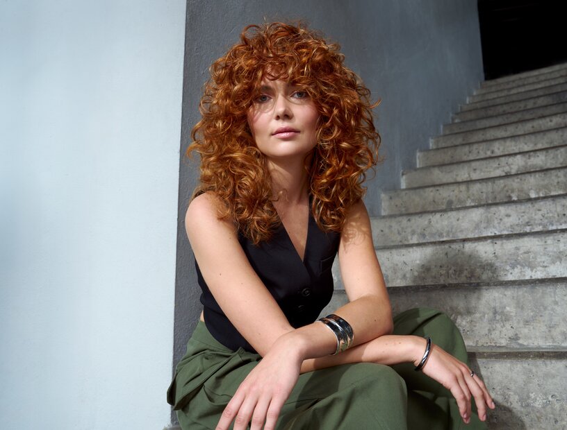 Porträt einer Frau mit voluminösen roten Locken, schwarzer ärmelloser Bluse und grüner Hose auf einer Betontreppe.