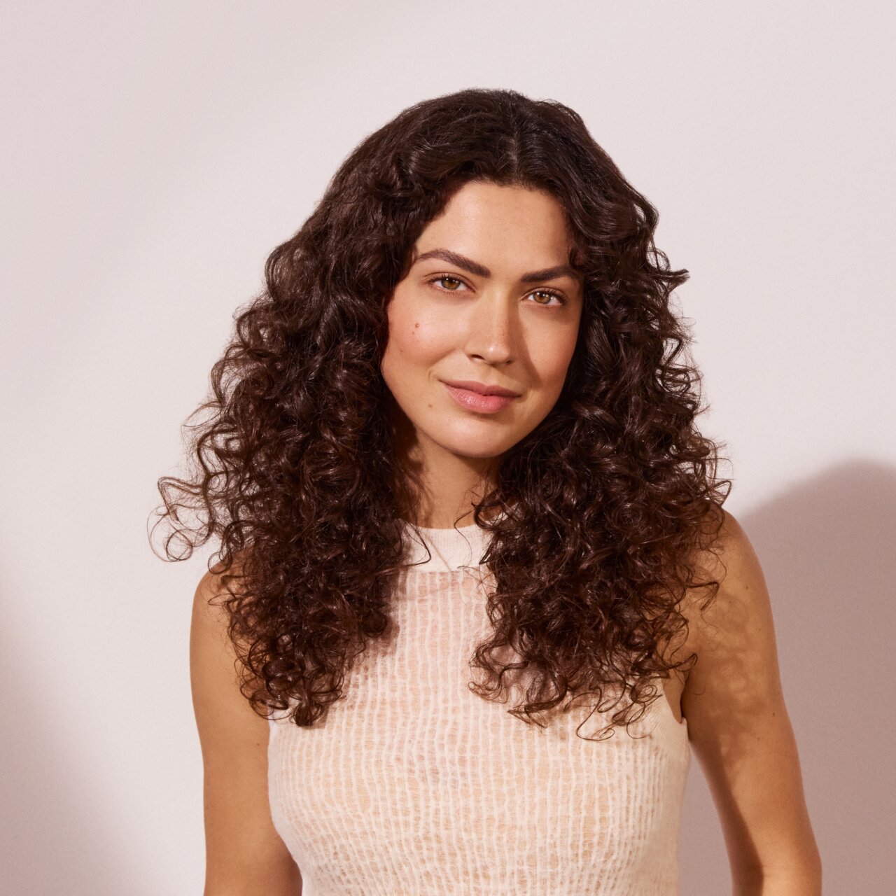  Frau mit langen dunkelbraunen Locken, die ein beigefarbenes Satinhemd trägt, vor einer hellen, rosefarbenen Wand. 