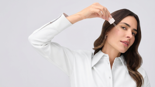  Frau mit weißer Bluse trägt mit einer Pipette ein transparentes Serum seitlich auf die Wange auf, vor hellgrauem Hintergrund.