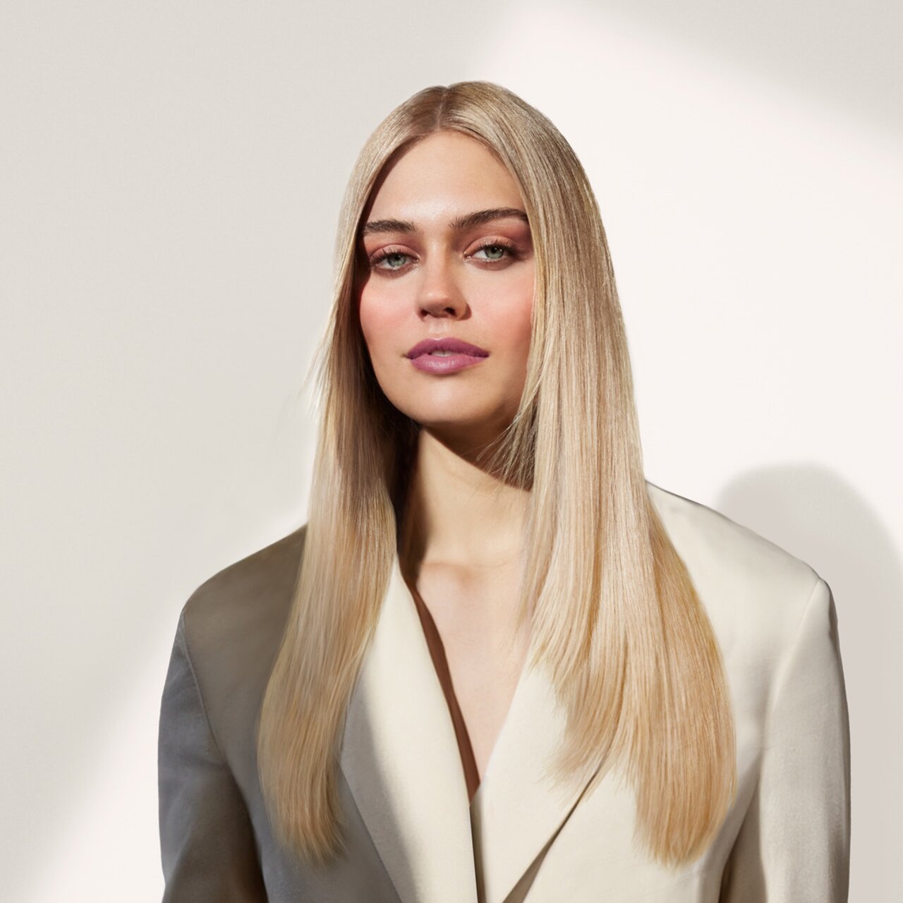 Eine junge Frau mit langen, glatten, blonden Haaren trägt einen eleganten, cremefarbenen Blazer vor einem schlichten, hellen Hintergrund.