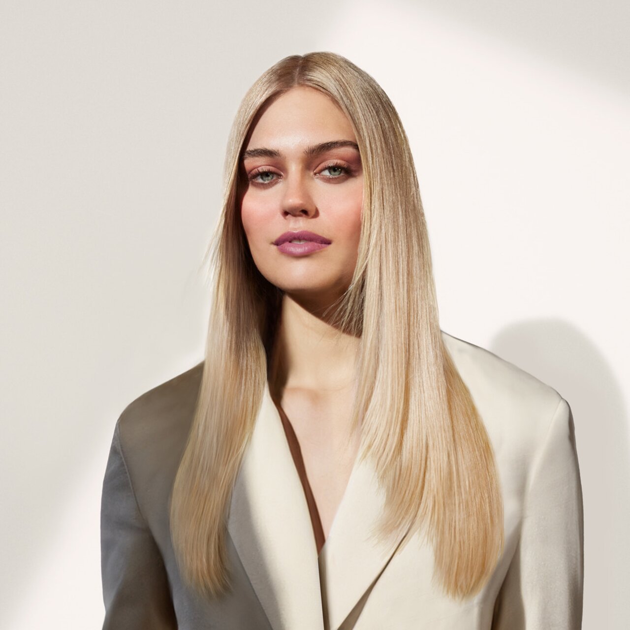 Sanfte Bronze- und Champagnertöne setzen beim Augen-Make-up im Frühjahr schimmernde Akzente. Eine junge Frau mit langen, glatten, blonden Haaren trägt einen eleganten, cremefarbenen Blazer vor einem schlichten, hellen Hintergrund.