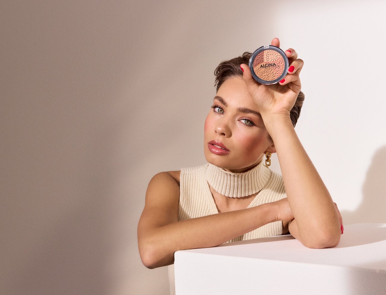 Junge schöne Frau mit geschminktem Gesicht stützt sich mit dem Arm auf einer Box ab und hält den ALCINA Powderblush in einer Hand, der für den Make-up-Trend Douyin Blush verwendet werden kann, da er vier verschiedene Farbnuancen enthält