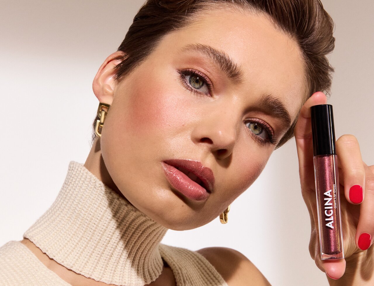 Eine schöne junge Frau mit zurück gekämmten Haaren und glitzerndem Lippenstift hält den Alcina Glam Lip Gloss in der Nuance Brown in den Händen und präsentiert den Make-up Trend Mocha Mousse Lips
