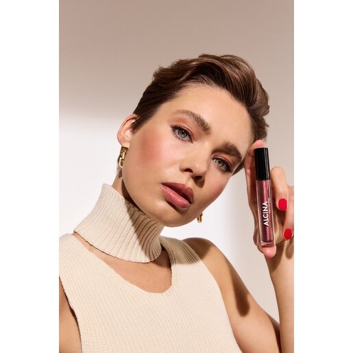 Eine schöne junge Frau mit zurück gekämmten Haaren und glitzerndem Lippenstift hält den Alcina Glam Lip Gloss in der Nuance Brown in den Händen und präsentiert den Make-up Trend Mocha Mousse Lips