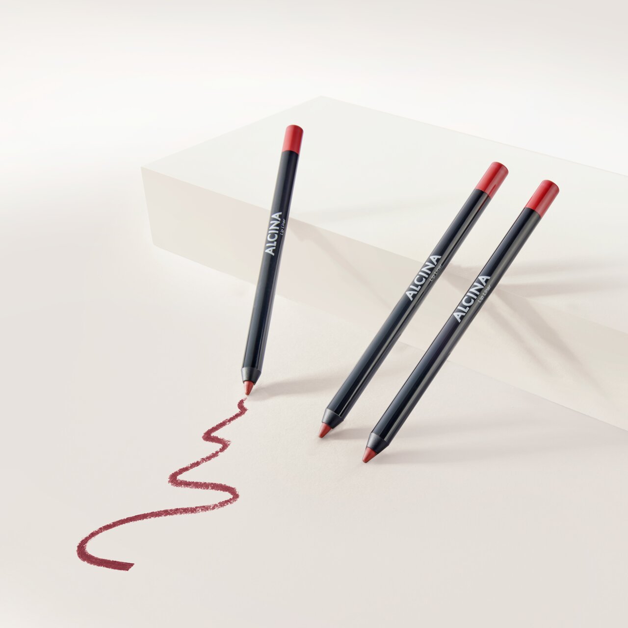ALCINA Lip Liner in dunklem Stiftdesign mit roter Mine, vor hellem Hintergrund arrangiert und mit geschwungener, aufgemalter Lippenkontur in sattem Rotton zur Veranschaulichung der Textur.