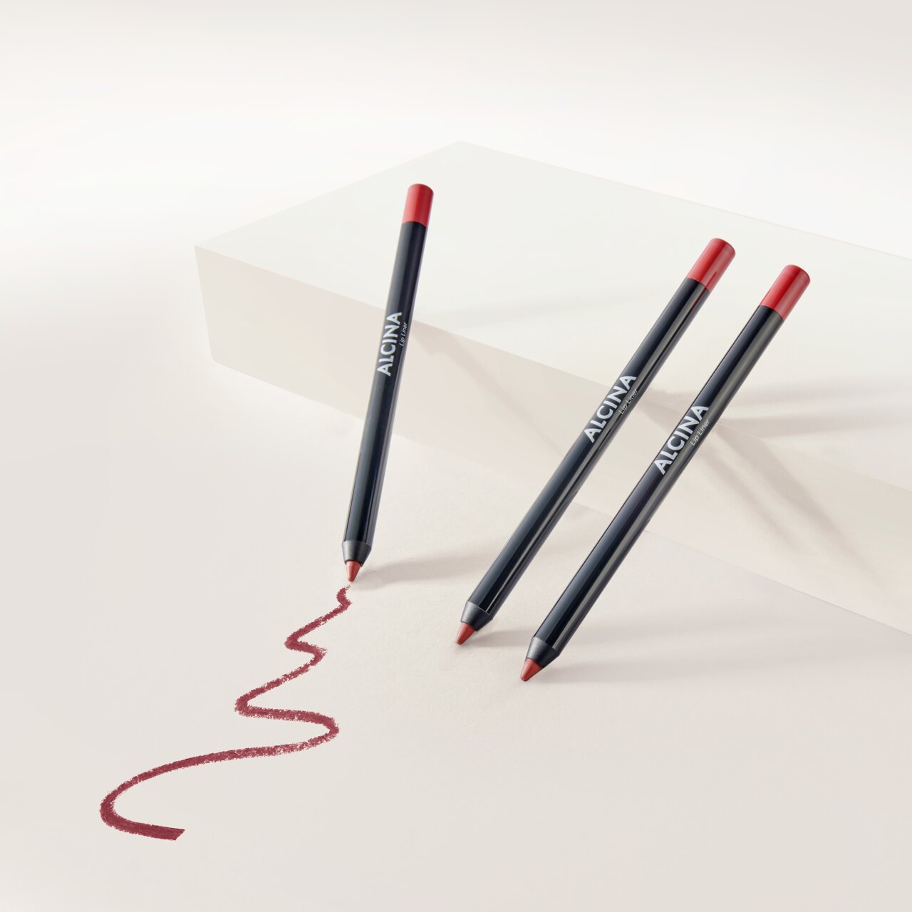 ALCINA Lip Liner in dunklem Stiftdesign mit roter Mine, vor hellem Hintergrund arrangiert und mit geschwungener, aufgemalter Lippenkontur in sattem Rotton zur Veranschaulichung der Textur.
