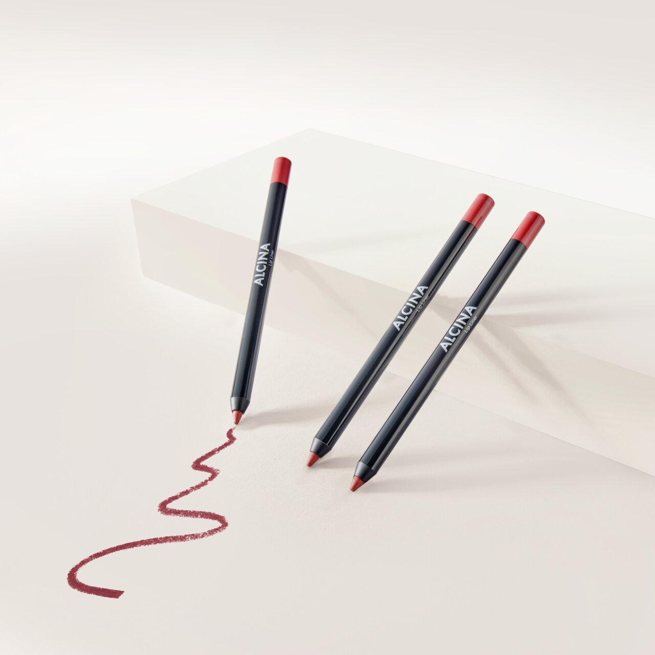 ALCINA Lip Liner in dunklem Stiftdesign mit roter Mine, vor hellem Hintergrund arrangiert und mit geschwungener, aufgemalter Lippenkontur in sattem Rotton zur Veranschaulichung der Textur.