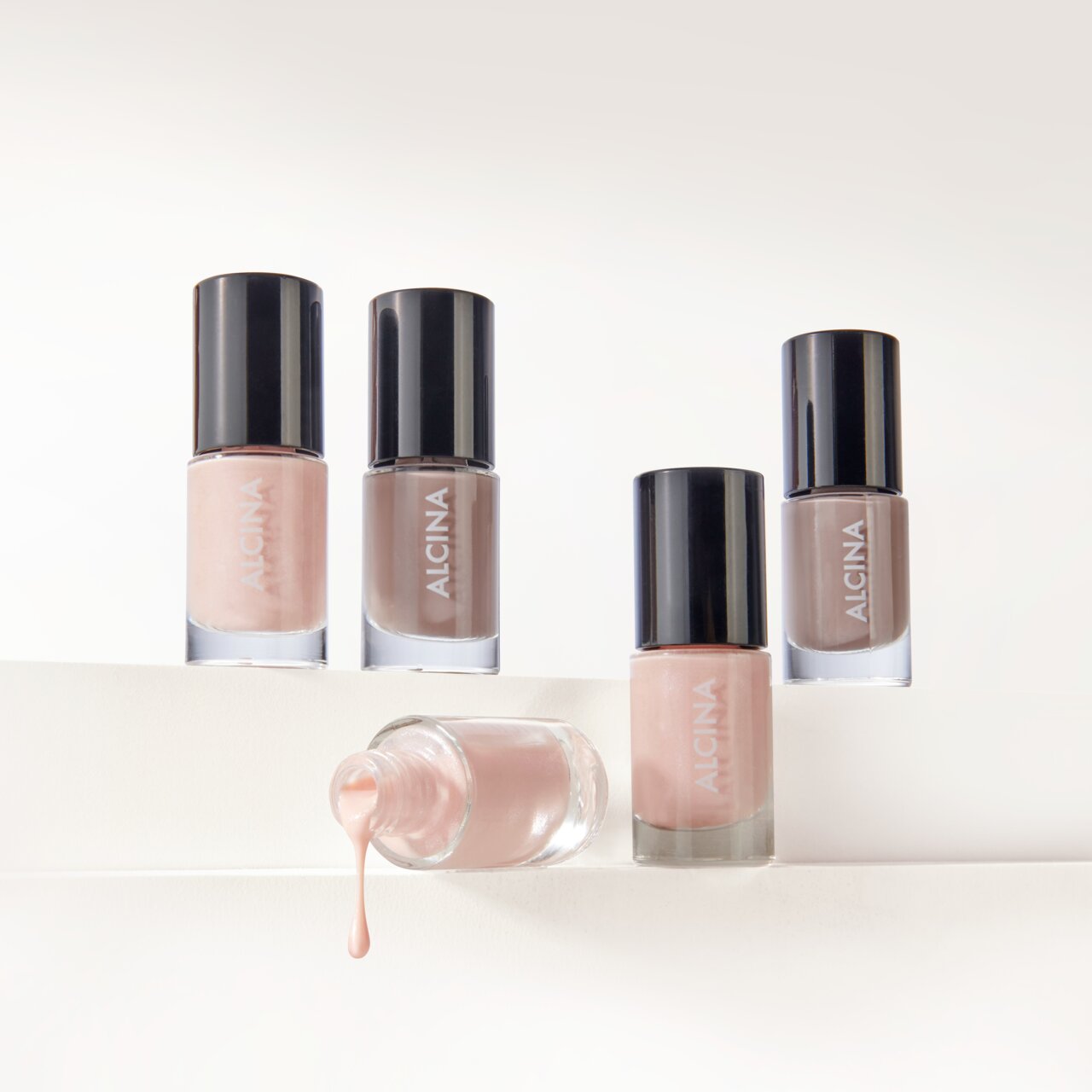 ALCINA Nail Color in den Nuancen rosy glam und elegance nude, mehrere Fläschchen mit glänzendem Finish vor hellem Hintergrund arrangiert, ein Flakon liegend mit herauslaufendem, zart roséfarbenem Nagellack.
