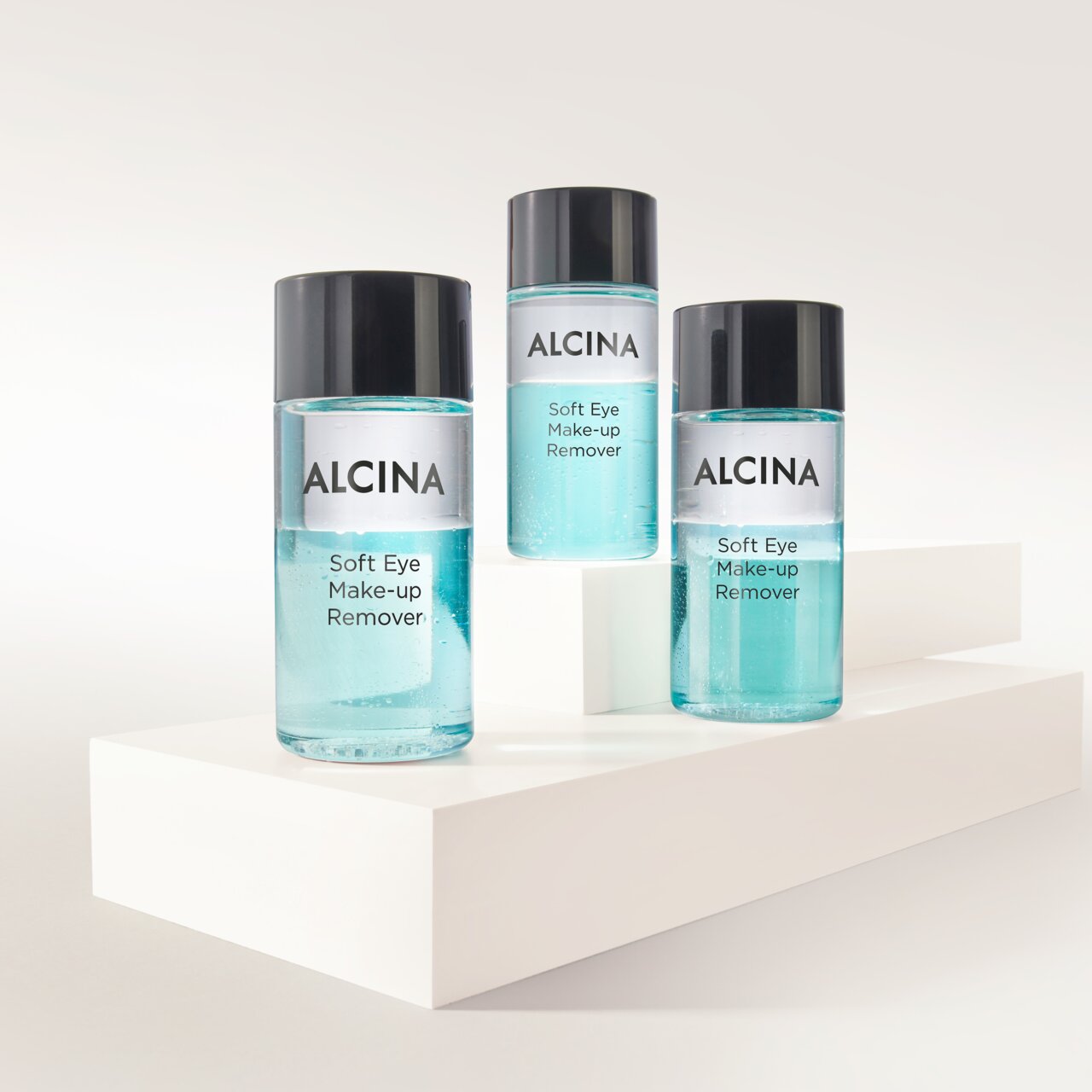 ALCINA Soft Eye Make-up Remover in drei transparenten Flaschen mit zweiphasiger, türkisfarbener Formulierung, auf weißen Podesten arrangiert für sanftes und gründliches Entfernen von Augen-Make-up.