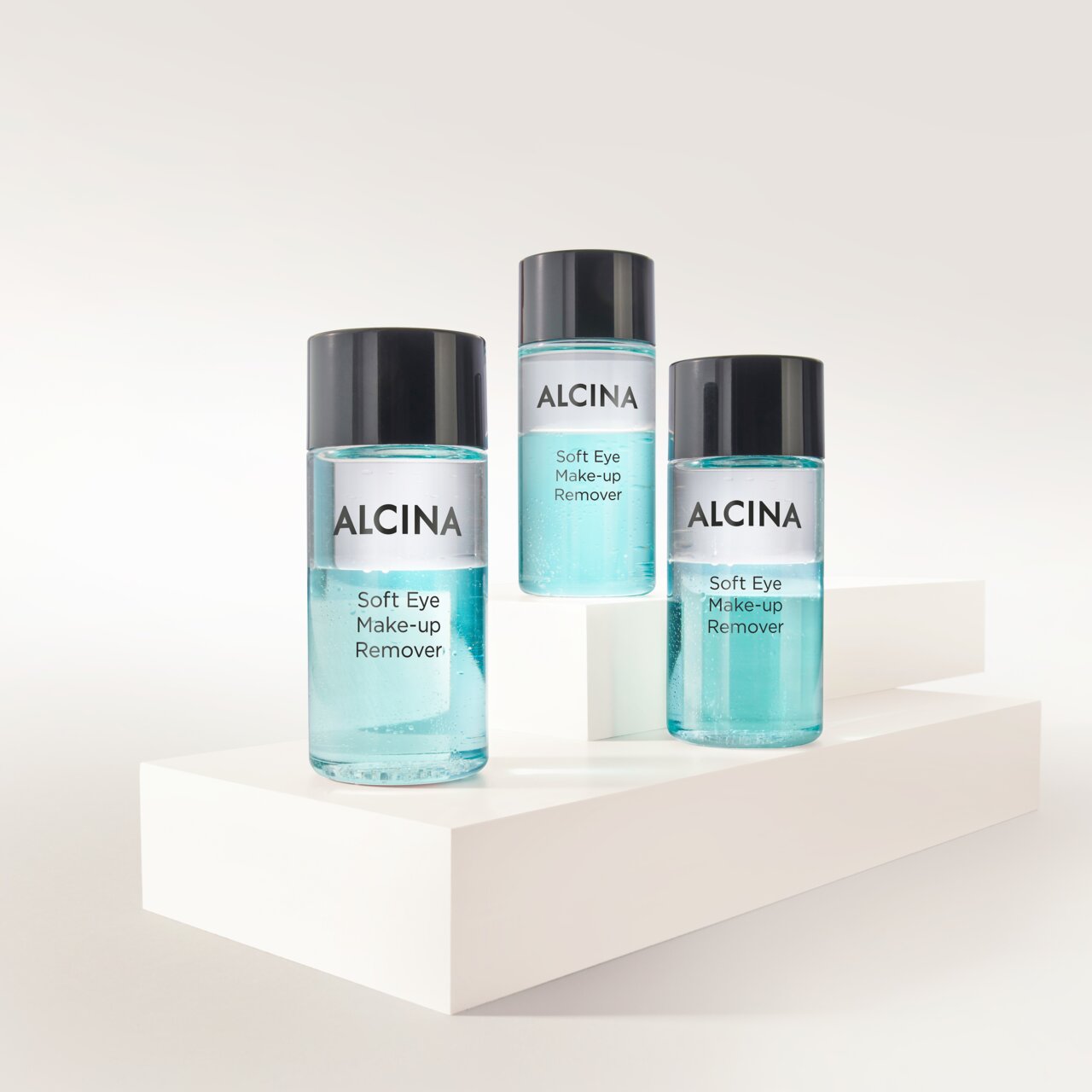 ALCINA Soft Eye Make-up Remover in drei transparenten Flaschen mit zweiphasiger, türkisfarbener Formulierung, auf weißen Podesten arrangiert für sanftes und gründliches Entfernen von Augen-Make-up.