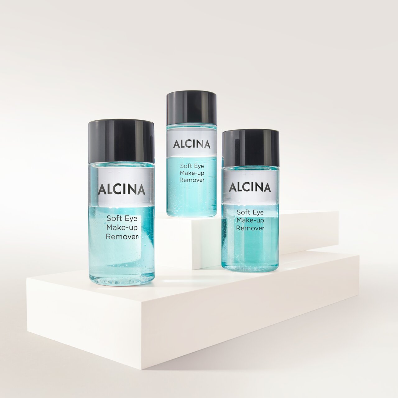 ALCINA Soft Eye Make-up Remover in drei transparenten Flaschen mit zweiphasiger, türkisfarbener Formulierung, auf weißen Podesten arrangiert für sanftes und gründliches Entfernen von Augen-Make-up.