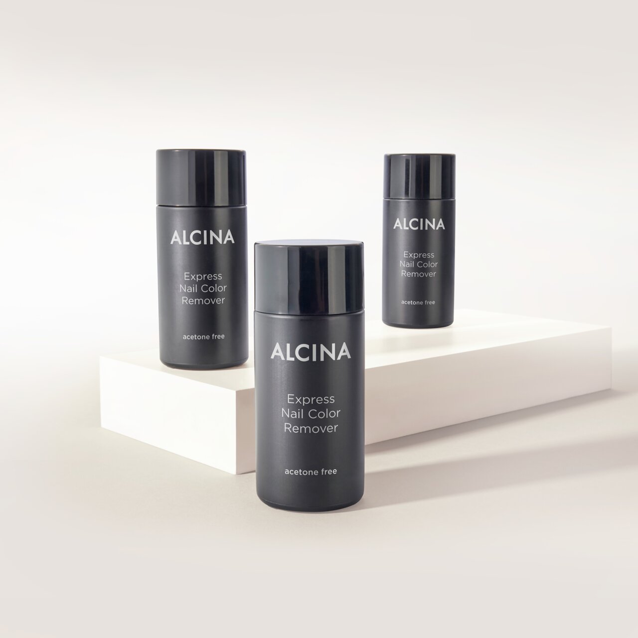 Drei schwarze Flaschen ALCINA Express Nail Color Remover stehen dekorativ auf hellen Podesten vor neutralem Hintergrund.