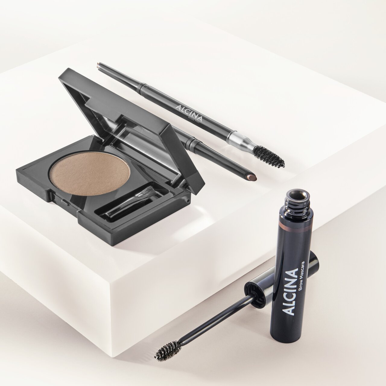 ALCINA Brow Powder, ALCINA Brow Pen und ALCINA Brow Mascara als Brow-Styling-Set dekorativ auf einem hellen Podest arrangiert.