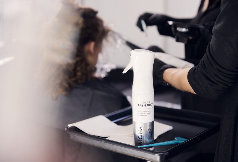 ALCINA A\CPlex Pre-Spray steht auf einem Friseurtablett im Vordergrund, während im Hintergrund eine Kundin mit lockigem Haar während einer Salonbehandlung betreut wird.