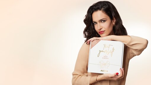 Schöne Frau mit rotem Lippenstift und rot lackierten Nägeln, die ein Alcina Weihnachten Beauty-Geschenkset in den Händen hält