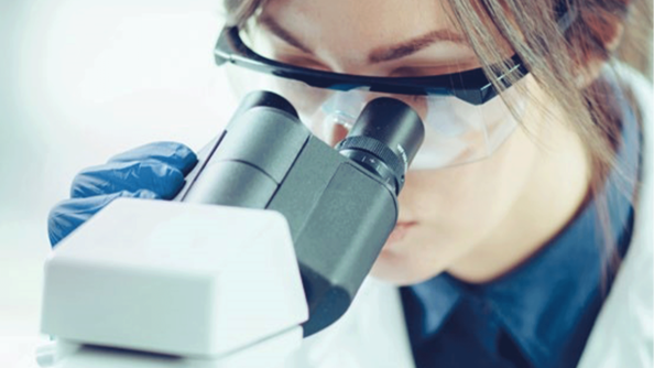 Nahaufnahme einer jungen Frau, die eine Laborbrille und blaue Latexhandschuhe trägt und durch ein Mikroskop sieht.