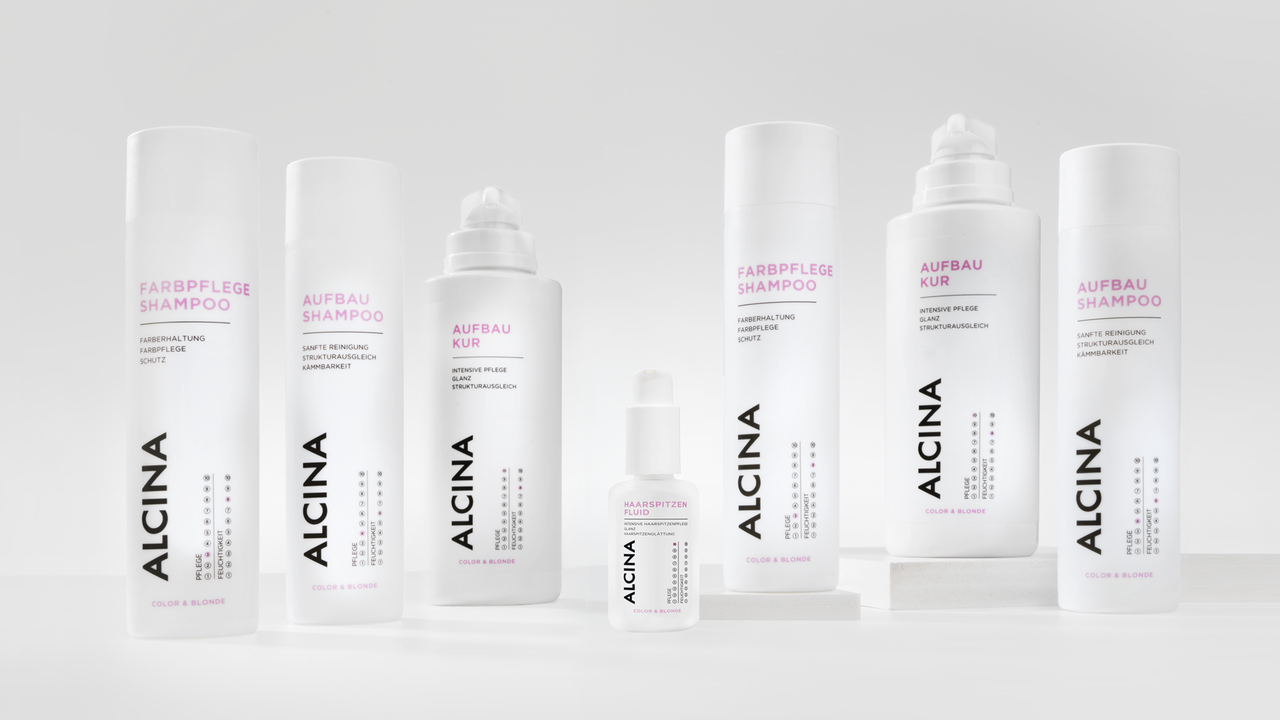 ALCINA Color und Blonde Haarpflegeserie mit Farbpflege-Shampoo, Aufbau-Kur, Aufbau-Shampoo und weiteren Produkten in weiß-pinkem, minimalistischem Design, übersichtlich auf heller Fläche arrangiert.