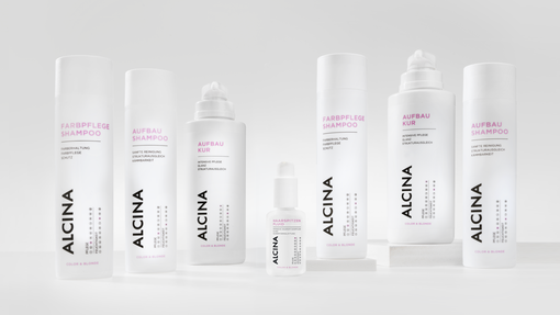ALCINA Color und Blonde Haarpflegeserie mit Farbpflege-Shampoo, Aufbau-Kur, Aufbau-Shampoo und weiteren Produkten in weiß-pinkem, minimalistischem Design, übersichtlich auf heller Fläche arrangiert.