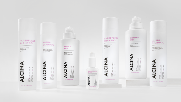 ALCINA Color und Blonde Haarpflegeserie mit Farbpflege-Shampoo, Aufbau-Kur, Aufbau-Shampoo und weiteren Produkten in weiß-pinkem, minimalistischem Design, übersichtlich auf heller Fläche arrangiert.