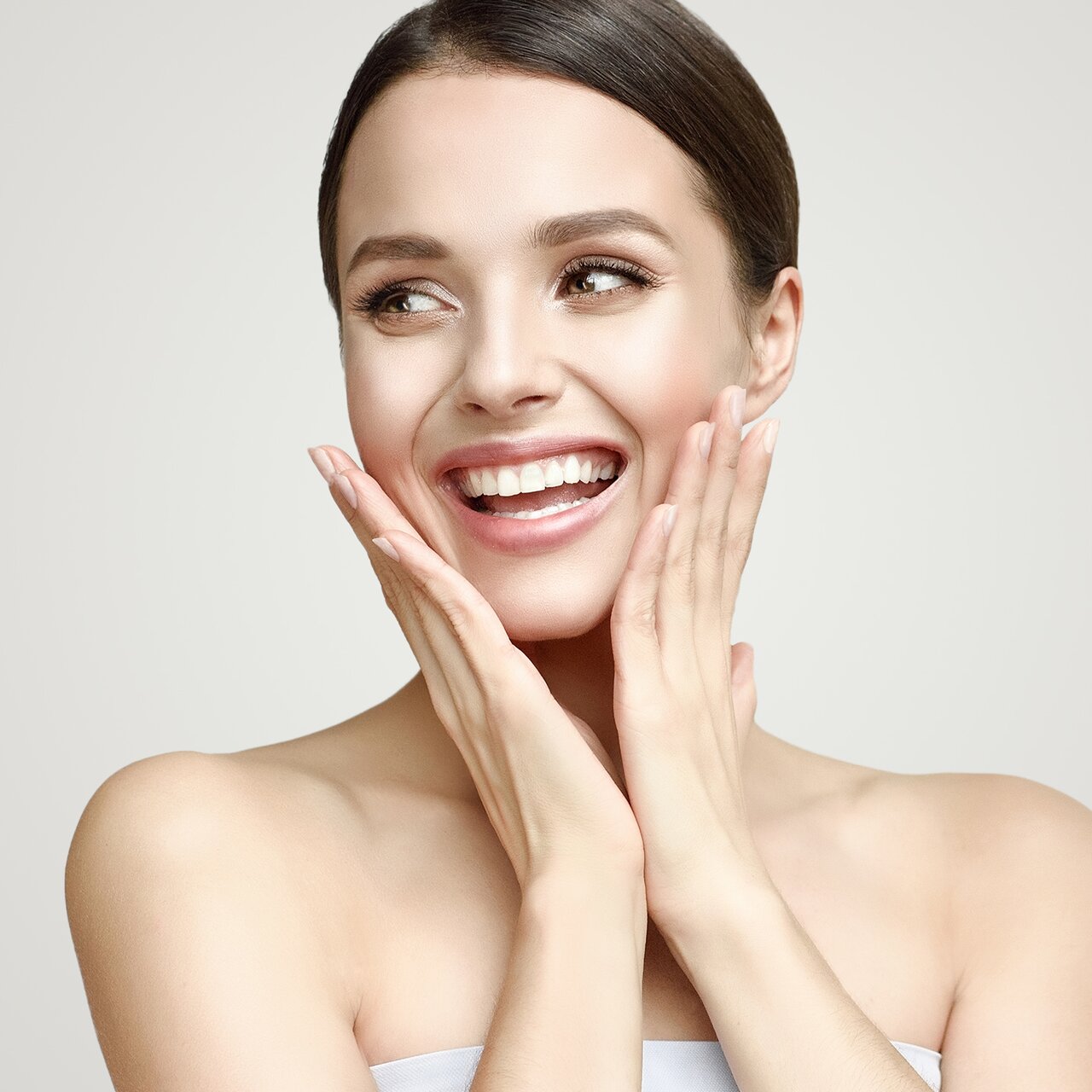 Lächelnde Frau mit schöner, gepflegter Haut, die zur Seite blickt und die Hände jeweils links und rechts an die Wangen legt