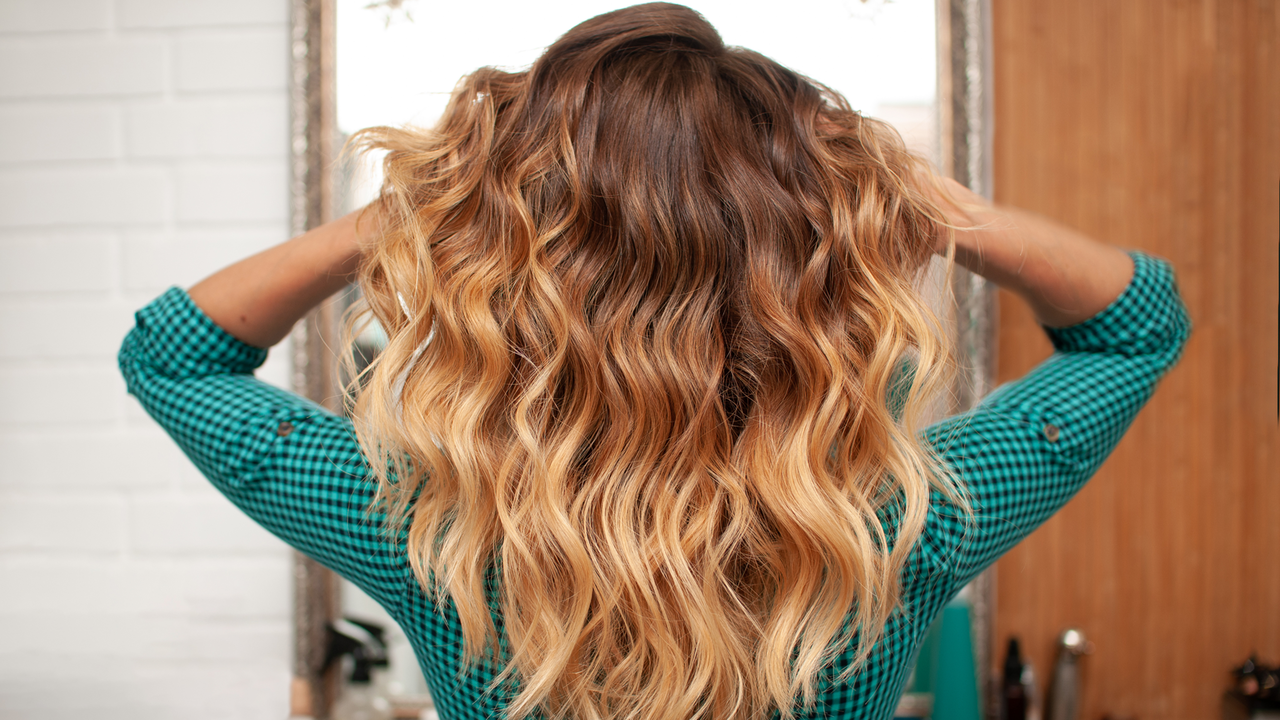 Ombre kann alles im Haar – vom natürlichen Übergang bis zu auffälligen Kontrasten Halbportrait einer Frau in Rückansicht, die sich mit beiden Händen in ihre leicht gewellte ombré-gefärbte Langhaarfrisur greift