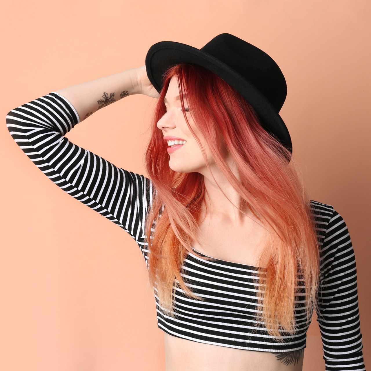 Auch mit bunten professionellen Haarfarben kann Ombré tolle Kontraste setzen Halbportrait einer Frau im kurzen Ringelpullover mit schwarzem Hut und langen, erdbeerblond gefärbten Haaren im Ombré-Stil, die sich mit einer Hand an den Hut fasst