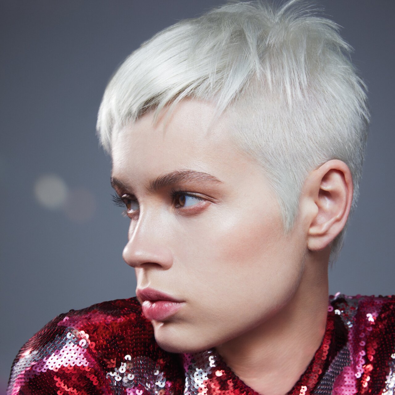 Porträt einer schönen jungen Frau mit der Trendfrisur 2024 Ultra Short Pixie in der Haarfarbe Platinblond, die den Kopf leicht zu Seite geneigt hat