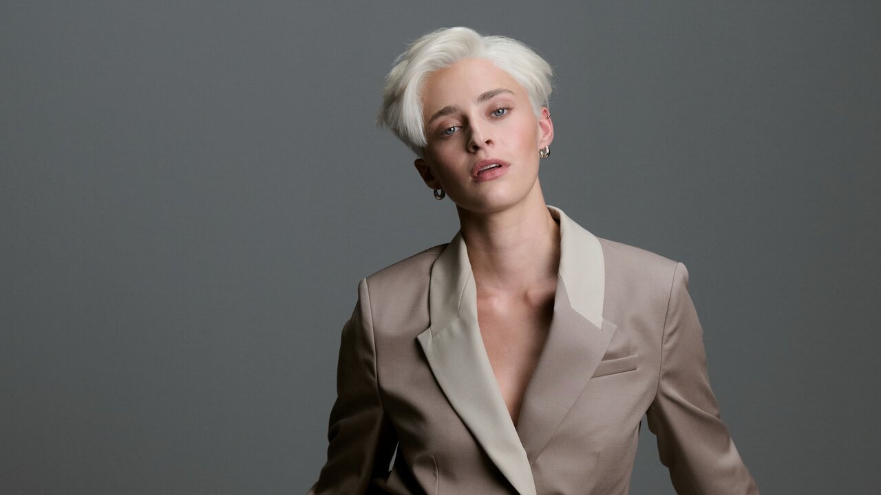 Androgyn gekleidete Frau mit kurz geschnittenen, platinblonden Haaren in einem zweifarbigen Blazer vor neutral grauem Hintergrund, der Oberkörper leicht nach vorne geneigt.