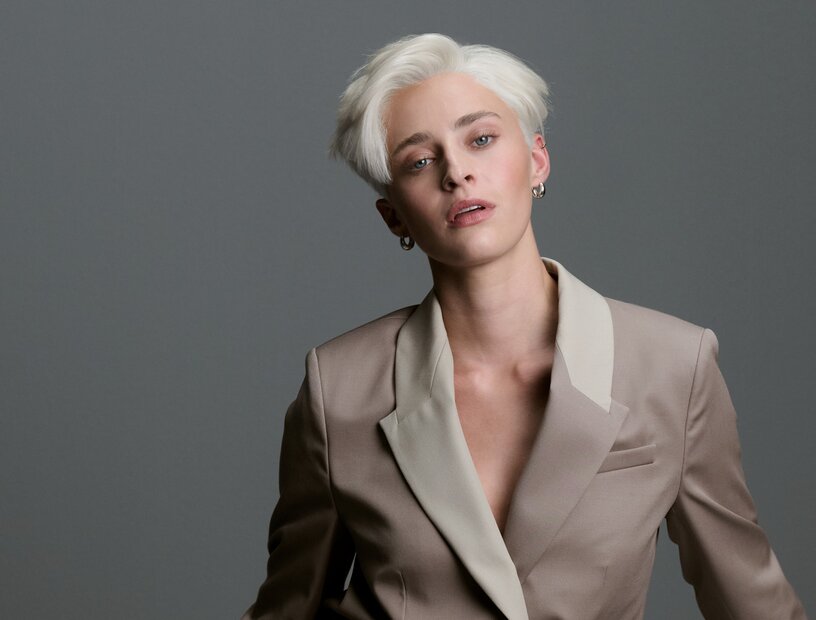 Androgyn gekleidete Frau mit kurz geschnittenen, platinblonden Haaren in einem zweifarbigen Blazer vor neutral grauem Hintergrund, der Oberkörper leicht nach vorne geneigt.