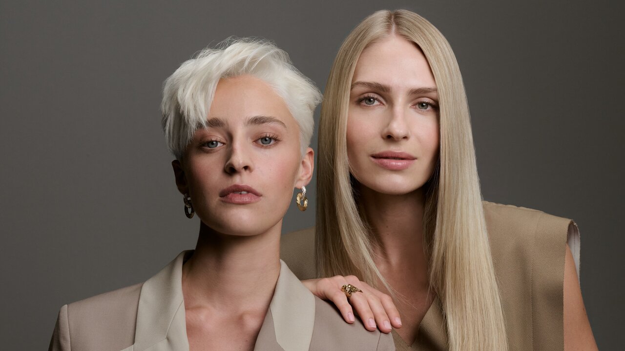 Zwei Frauen mit kontrastierenden Blondtönen – links ein kühles, platinblondes Kurzhaar, rechts ein warmes, goldblondes Langhaar – posieren in beigefarbenen Outfits vor grauem Hintergrund