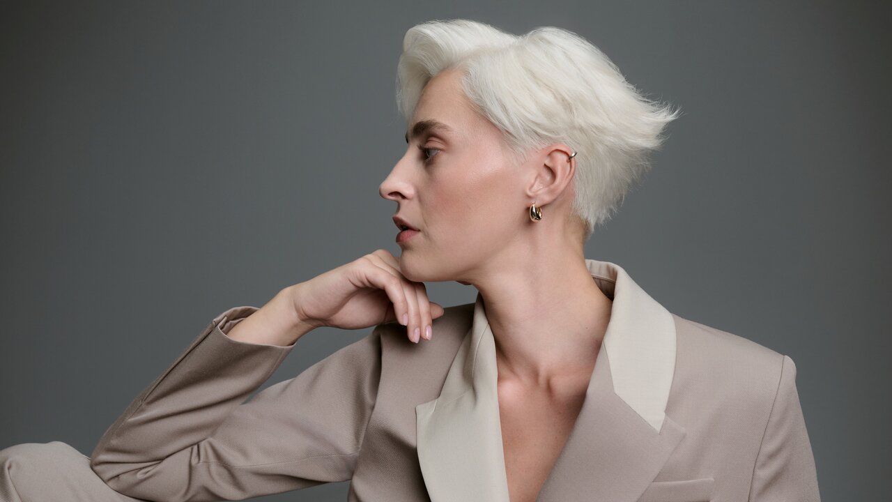 Frau mit kurzem, kühlem Platinblond und strukturiertem Seitenscheitel, lässig in einem beigefarbenen Blazer vor grauem Hintergrund sitzend.