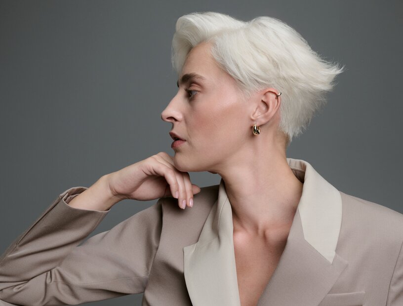 Frau mit kurzem, kühlem Platinblond und strukturiertem Seitenscheitel, lässig in einem beigefarbenen Blazer vor grauem Hintergrund sitzend.