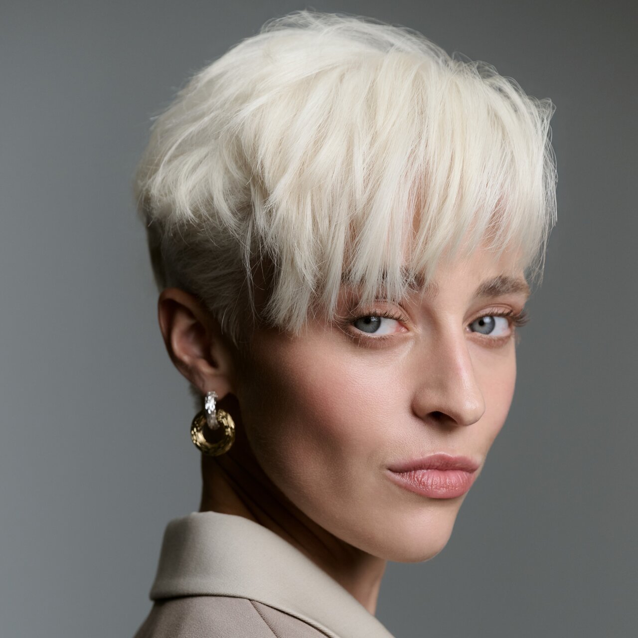 Kurz geschnittenes, kühles Platinblond mit weicher Struktur, seitlich von hinten aufgenommen, kombiniert mit goldenen Creolen vor neutral grauem Hintergrund.