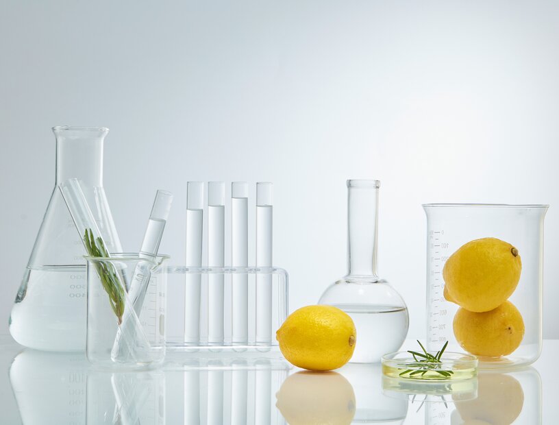 Reagenzgläser, Kolben und Messbecher, die mit klarer Flüssigkeit und Zitronen gefüllt sind als Sinnbild für den Einsatz von Vitamin C in der Hautpflege